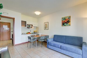 A living room with a blue sofa, a dining table with chairs, a kitchen area with cabinets and a microwave, and a door with a notice.