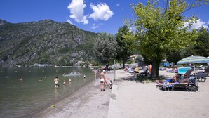 A beach with people swimming and sunbathing, a mountain in the background, and a tree providing shade. - Porto Letizia P11, Porlezza, Italy (Porlezza)