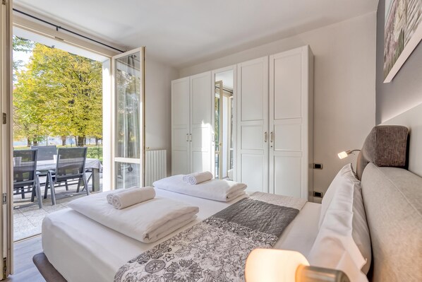 A hotel room with a bed, a seating area with a table and chairs, and a view of trees outside. - Porto Letizia Iris 2, Porlezza, Italy (Porlezza)