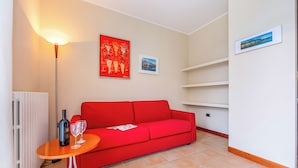 Ein Wohnzimmer mit einem roten Sofa, einem kleinen Holztisch mit einer Flasche und Gläsern, einer Stehlampe und zwei gerahmten Bildern an der Wand.