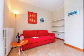 Een woonkamer met een rode bank, een klein houten tafeltje met een fles en glazen, een vloerlamp en twee ingekaderde foto's aan de muur.
