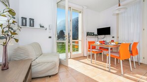 En moderne stue med sofa, spisebord med orange stole, fjernsyn og en skydedør til en balkon med udsigt over et bjerg.