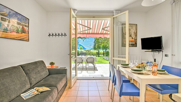 A living room with a sofa, a dining table with chairs, a TV, and a view of the outdoors. - Porto Letizia Begonia 4, Porlezza, Italy (Porlezza)