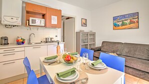 A modern kitchen with white cabinets, a microwave, and a sink. There is a dining table set with plates, glasses, and a bowl of fruit. A gray sofa and a painting of vintage cars are in the living area. - Porto Letizia Begonia 4, Porlezza, Italy (Porlezza)