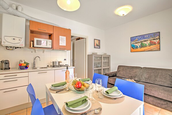 A modern kitchen with white cabinets, a microwave, and a sink. There is a dining table set with plates, glasses, and a bowl of fruit. A gray sofa and a painting of vintage cars are in the living area.