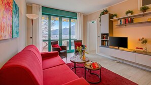 A modern living room with a red sofa, a small round table with a book and glasses, a TV on a white cabinet, and a view of mountains through large windows.