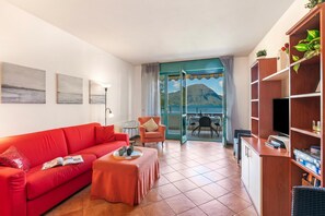 A living room with a red sofa, a small table, a chair, a TV, and a bookshelf. There is a balcony with a view of mountains. - Porto Letizia P13 B1, Porlezza, Italy (Porlezza)