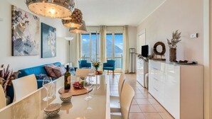 A modern living room with a sofa, dining table, and chairs. There are paintings on the wall, a balcony with a view, and a kitchen area with cabinets and a countertop. - Porto Letizia T12 B1, Porlezza, Italy (Porlezza)