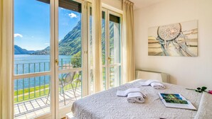 A hotel room with a large window, a bed with neatly folded towels, a framed picture on the wall, and a balcony with a chair. - Porto Letizia T12 B1, Porlezza, Italy (Porlezza)