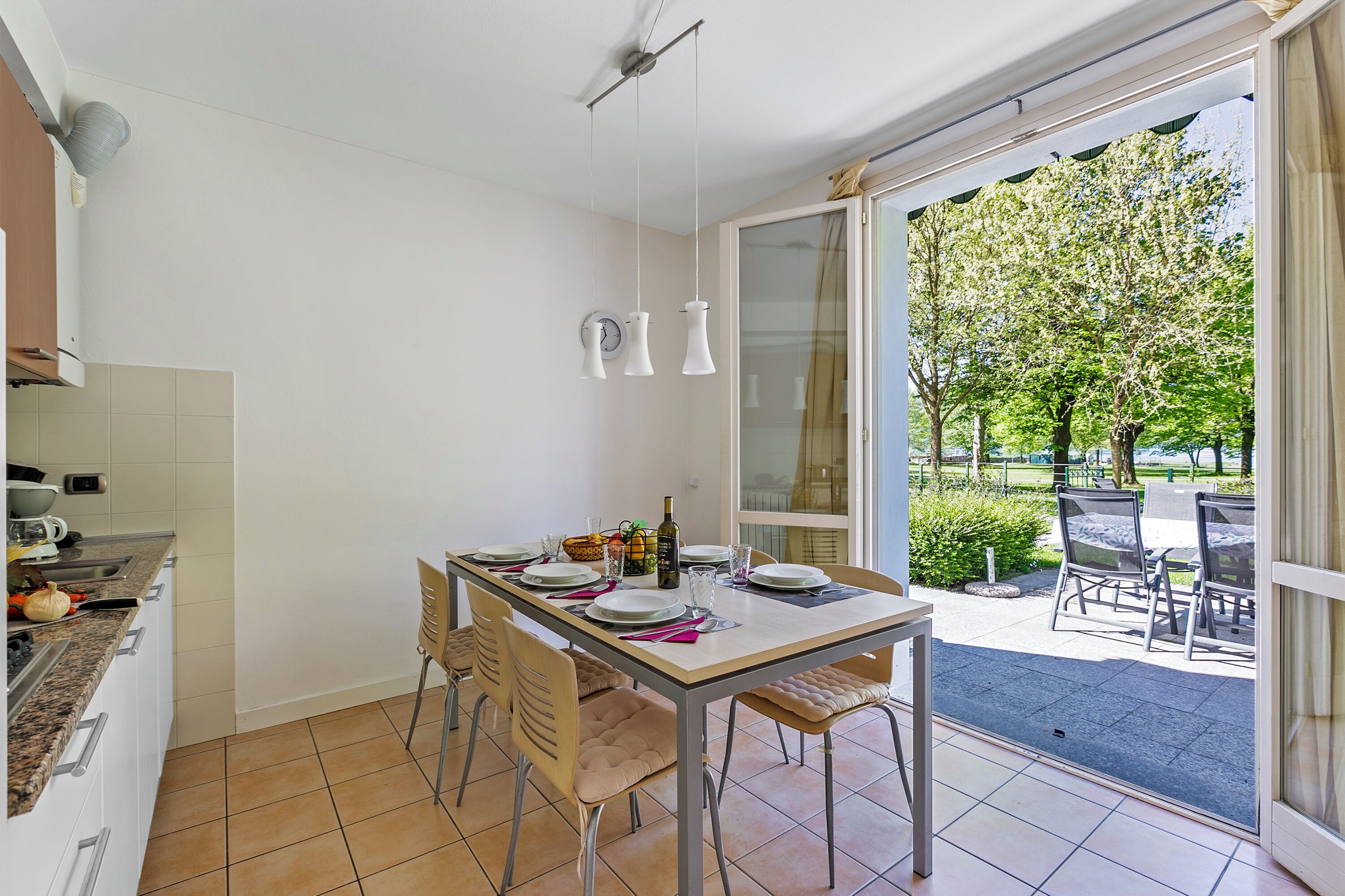 Ein Essbereich mit Tisch für vier Personen, eine Küche mit Herd und Spüle sowie Blick auf eine Terrasse mit Sitzgelegenheiten.