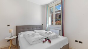 A neatly made bed with white linens and pillows, a bedside table with a lamp, and a window with curtains. - Porto Letizia Iris 1, Porlezza, Italy (Porlezza)
