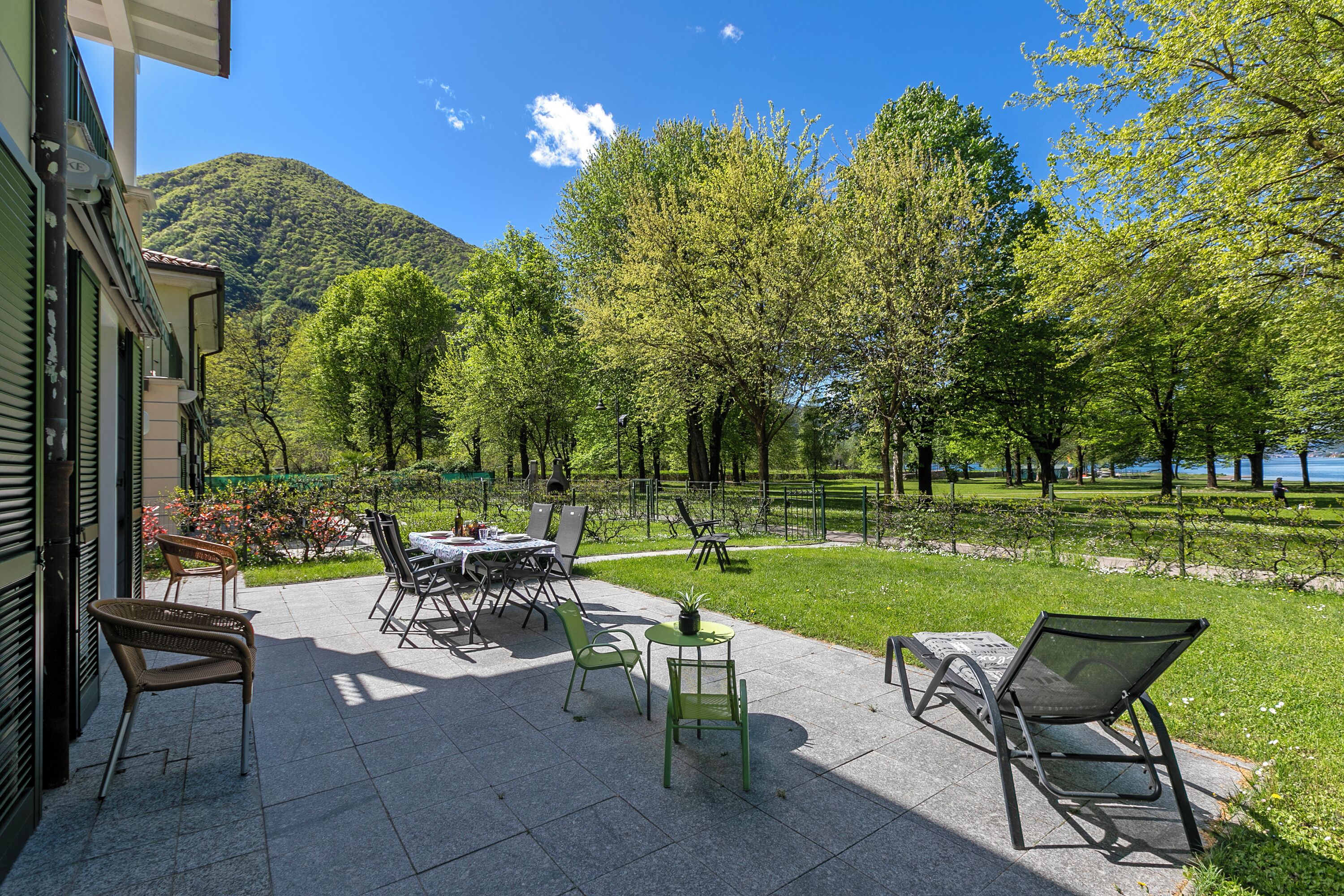 An outdoor patio with tables and chairs, a small round table with a plant, and a view of a lake and mountains in the background.