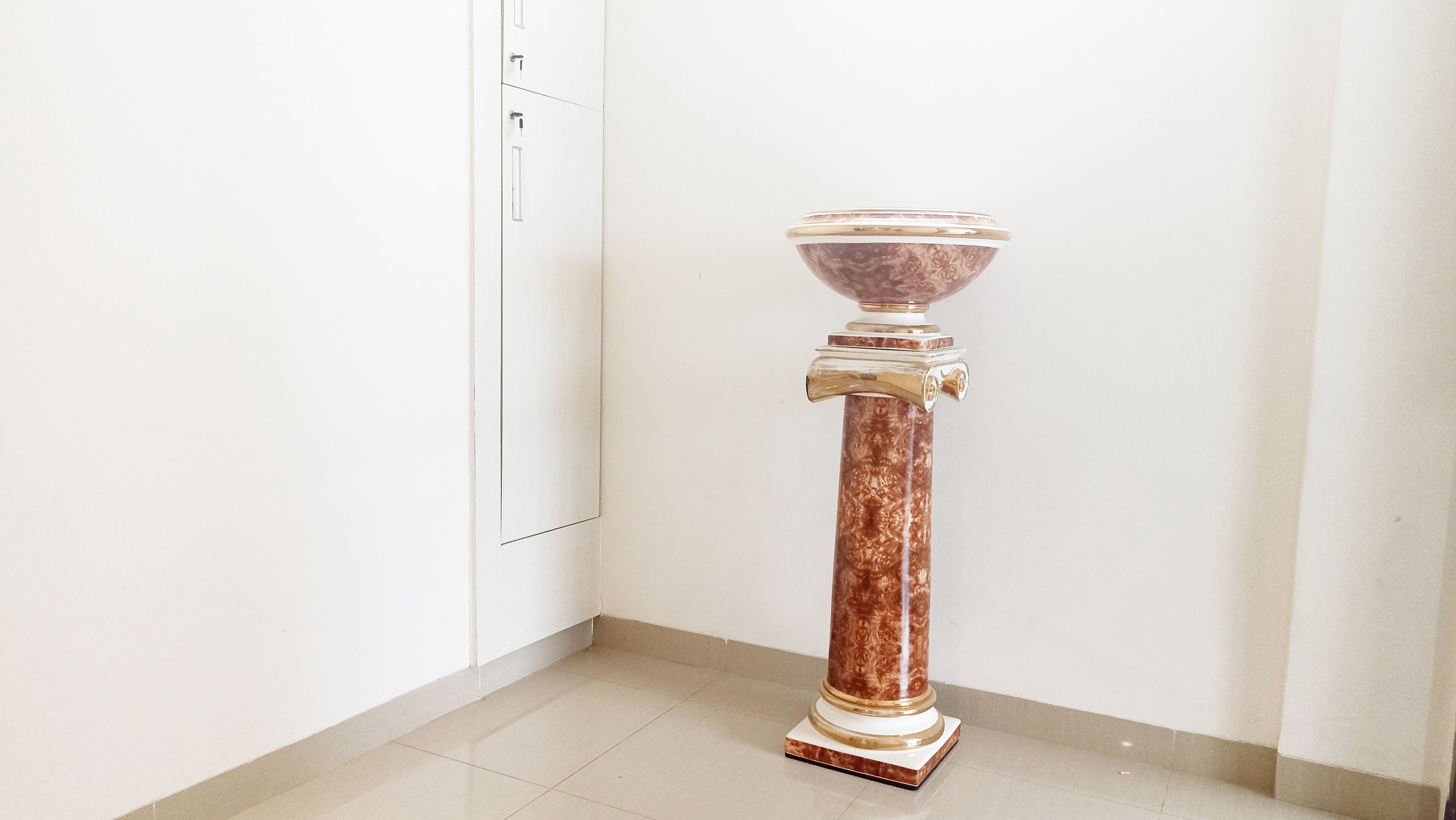 A marble pedestal table with a bowl on top, positioned in a corner of a room with white walls and a tiled floor.