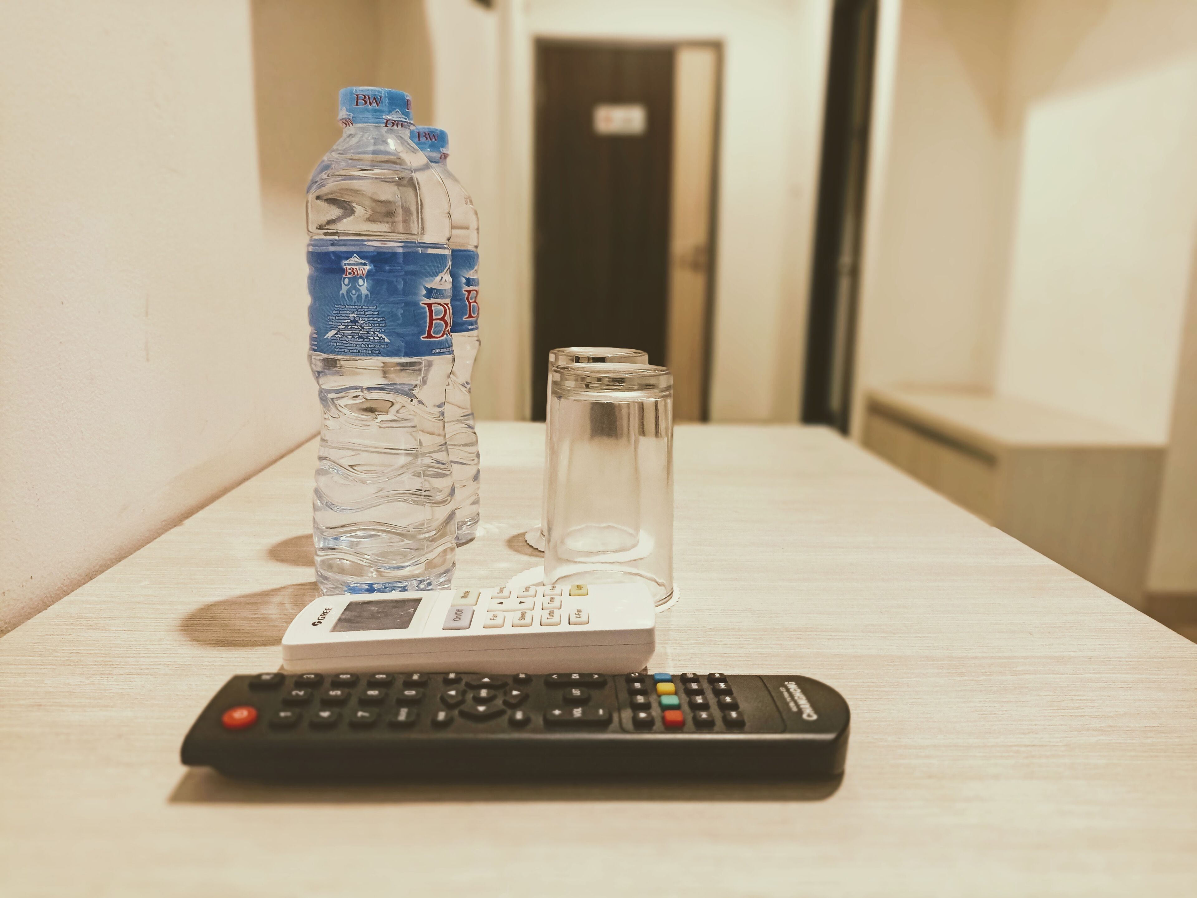 A remote control, a glass of water, and a bottle on a wooden table in a hotel room.