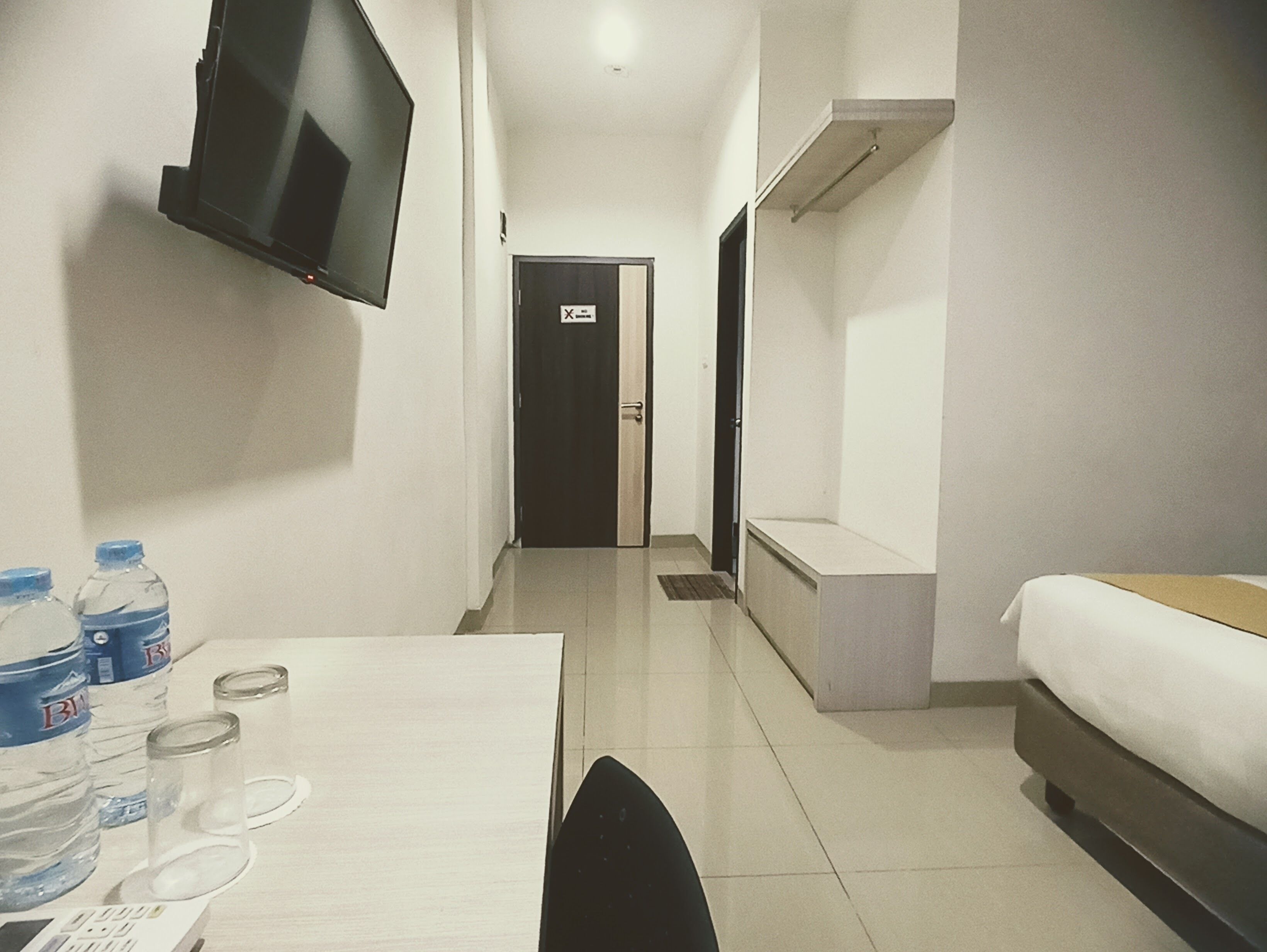 Quarto de hotel com uma cama, televisão fixada na parede, escrivaninha com cadeira e dispensador de água.