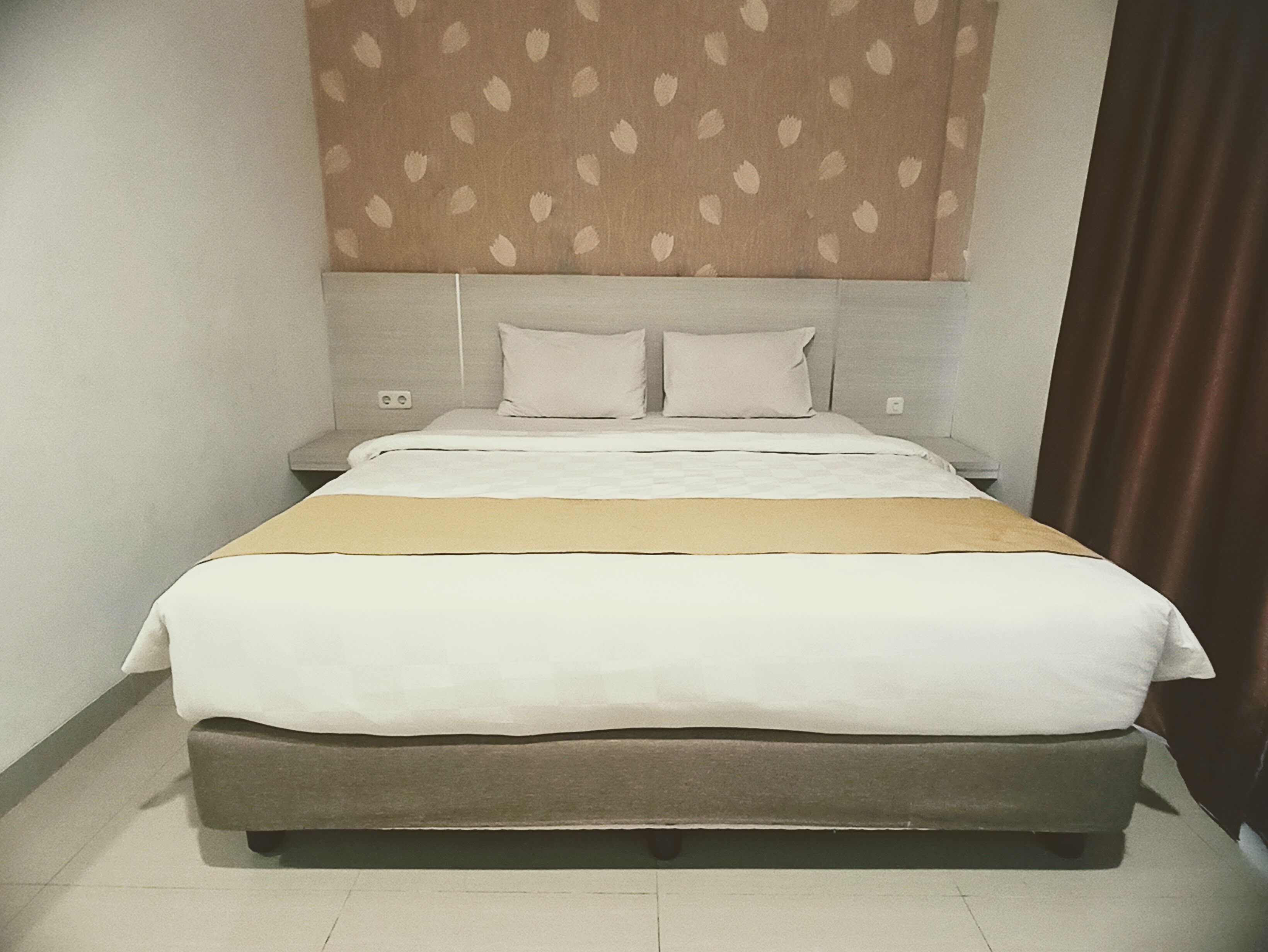 A neatly made bed with a white and beige bedspread, a grey headboard, and a patterned wall above.