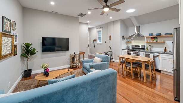A modern living room with a blue sofa, a wooden dining table, and a kitchen area with white cabinets and a stainless steel refrigerator. - / Luxury Modern Home in Oakland with AC & WFH / (Oakland)