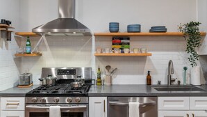 A modern kitchen with stainless steel appliances, wooden shelves, and a variety of kitchenware. - / Luxury Modern Home in Oakland with AC & WFH / (Oakland)