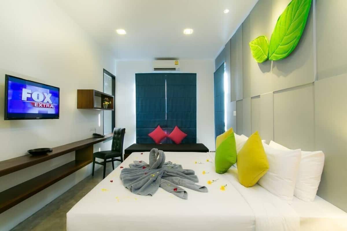 A hotel room with a TV displaying FOX EXTRA, a bed with white and colorful pillows, a desk with a laptop, and a wall-mounted air conditioner.