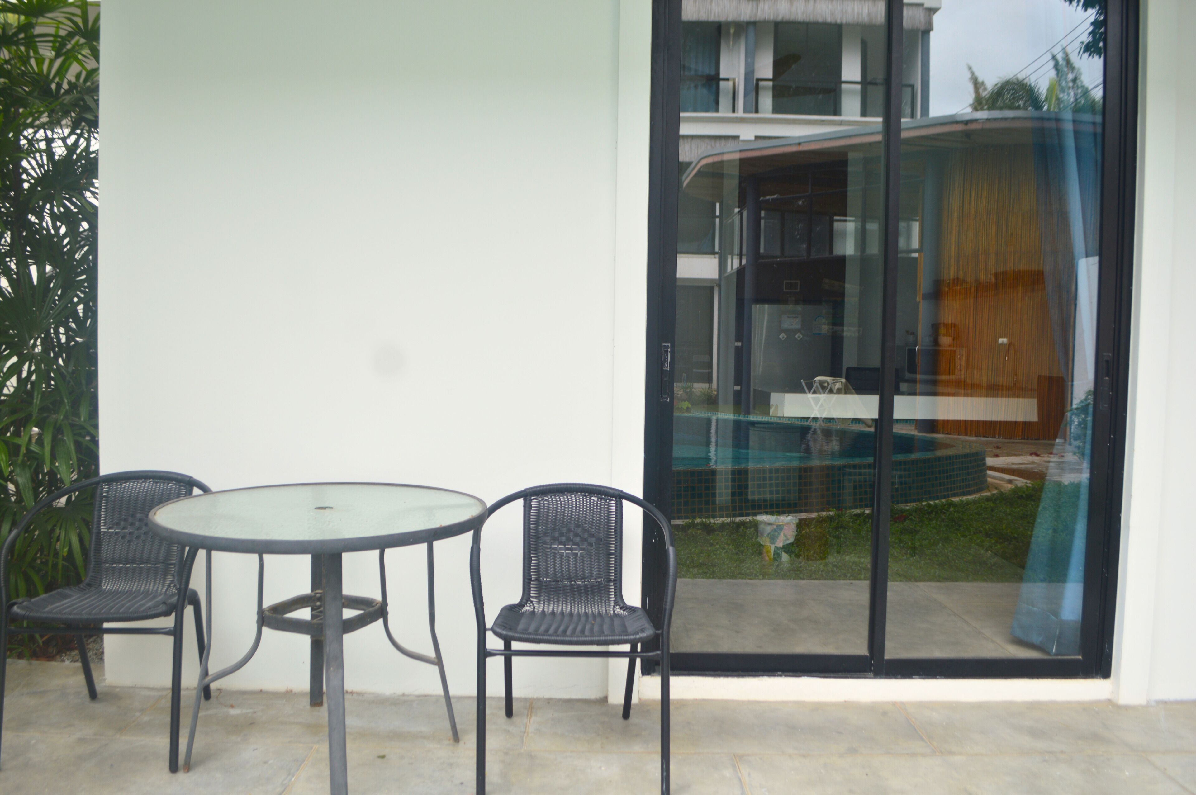 A patio with a glass table and two chairs, a sliding glass door, and a view of a pool and building.