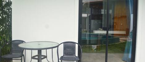 A patio with a glass table and two chairs, a sliding glass door, and a view of a pool and building.