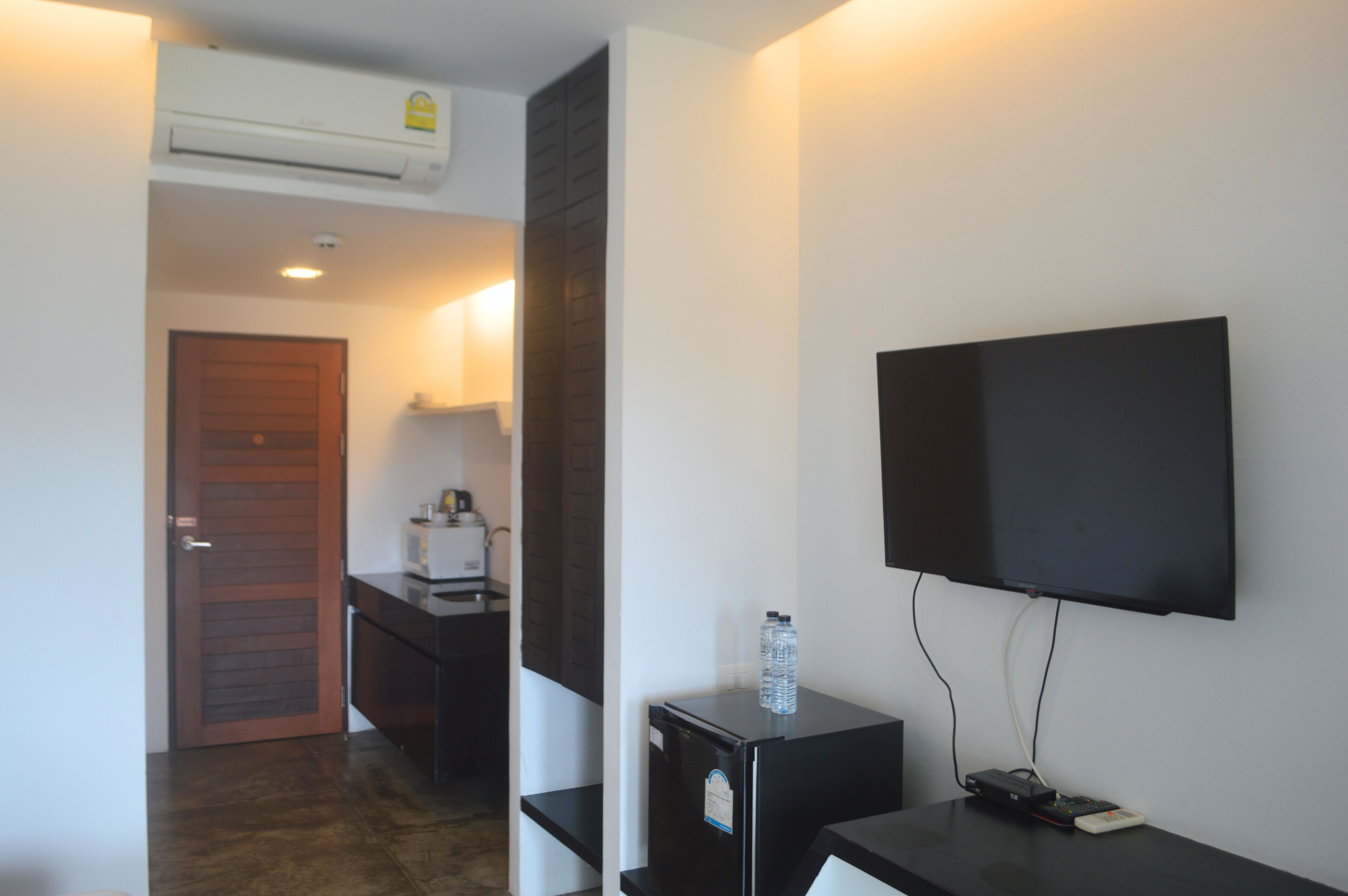 A modern hotel room with a flat-screen TV mounted on the wall, a small cabinet with a water bottle, and a door leading to the hallway.