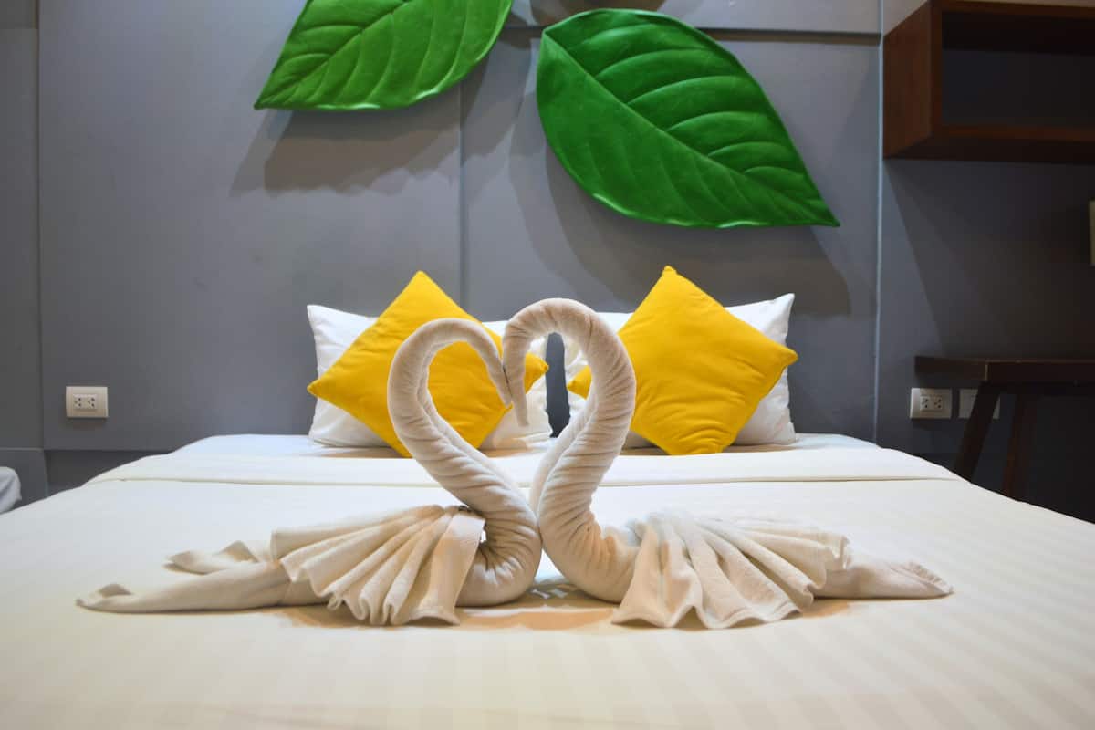 A hotel room with a bed featuring a towel art arrangement in the shape of a heart, yellow and white pillows, and green leaf decorations on the wall.