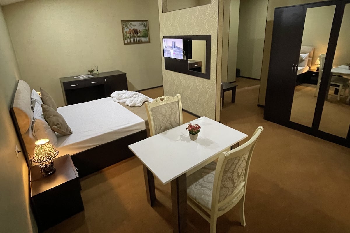Une chambre d’hôtel comprenant un lit, une table de chevet avec une lampe, une petite table avec un vase de fleurs, une chaise, un bureau et une armoire.