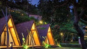 Una serie de cabañas tipo A, con forma triangular y cálida iluminación, rodeadas de abundante vegetación y un sendero de piedra.
