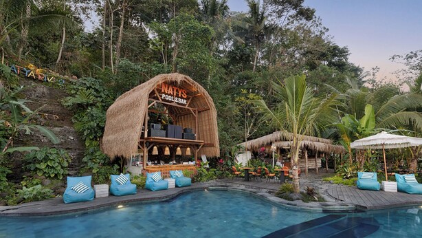 Zona de piscina con un bar de techo de paja llamado "NATYS POOL & BAR", rodeada de exuberante follaje y plantas tropicales.