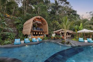 Zona de piscina con un bar de techo de paja llamado "NATYS POOL & BAR", rodeada de exuberante follaje y plantas tropicales.