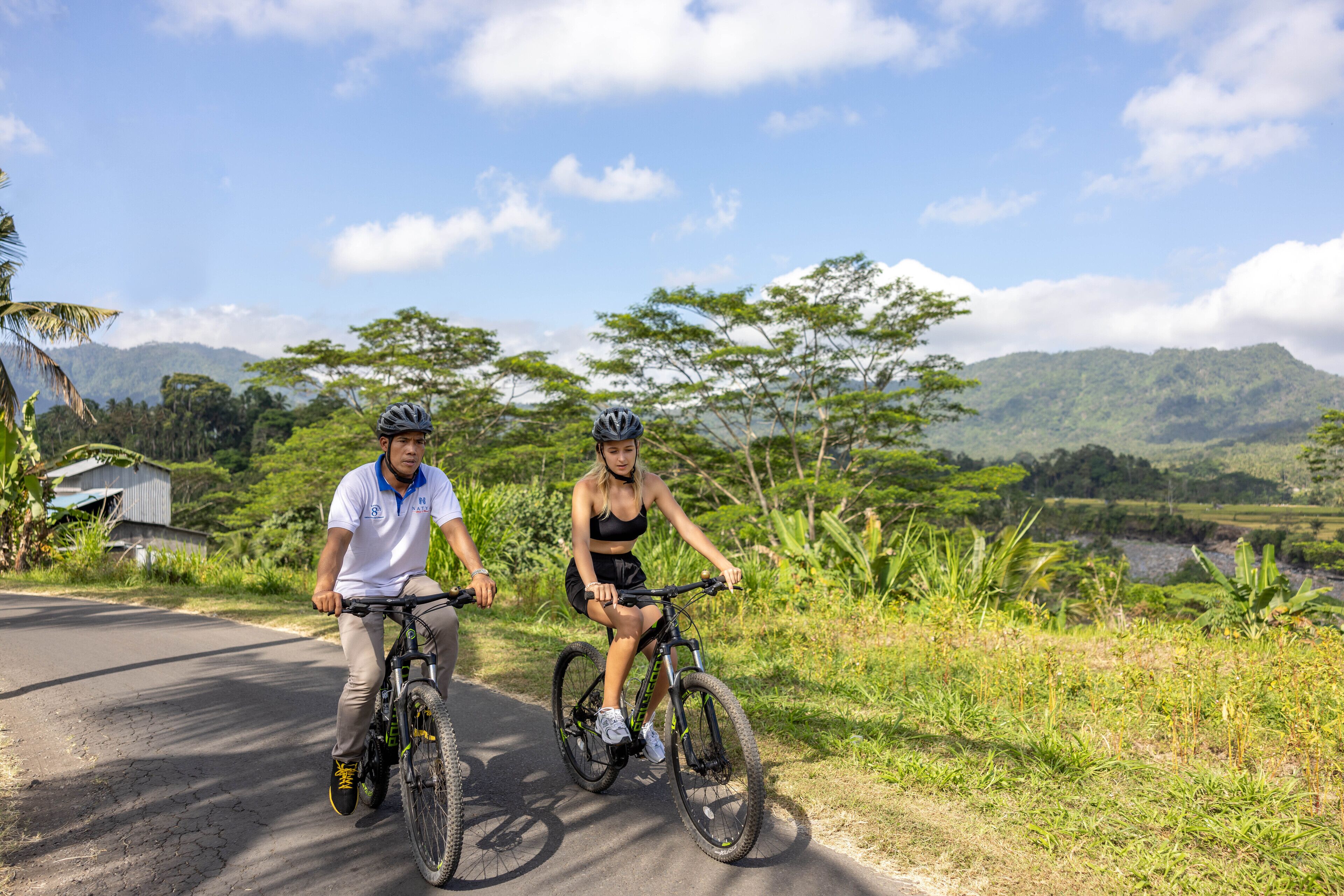 Two people riding bicycles on a paved road with greenery and hills in the background.