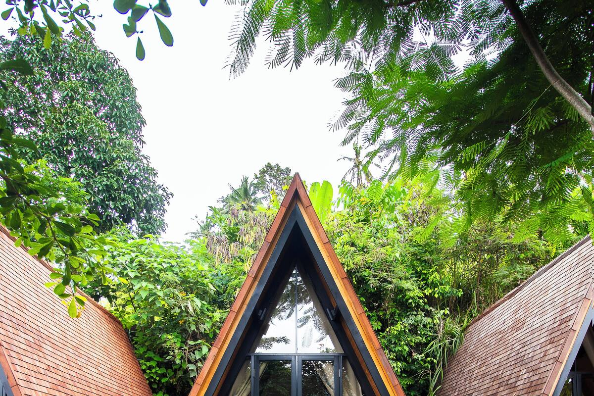 A triangular wooden structure with a glass door, surrounded by lush greenery and a grassy area with steps leading to a seating area.