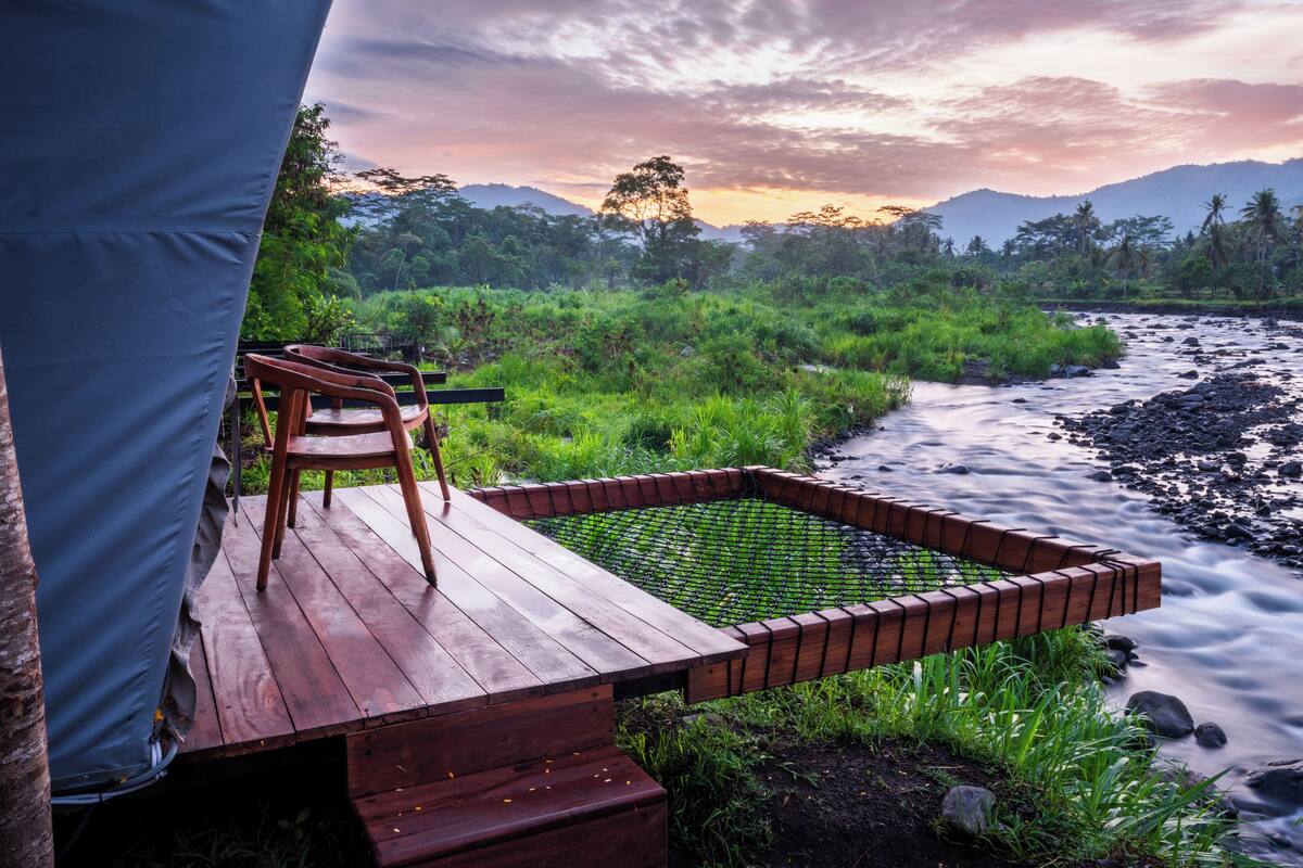 A wooden deck with a bench overlooking a river, surrounded by lush greenery and hills in the background.