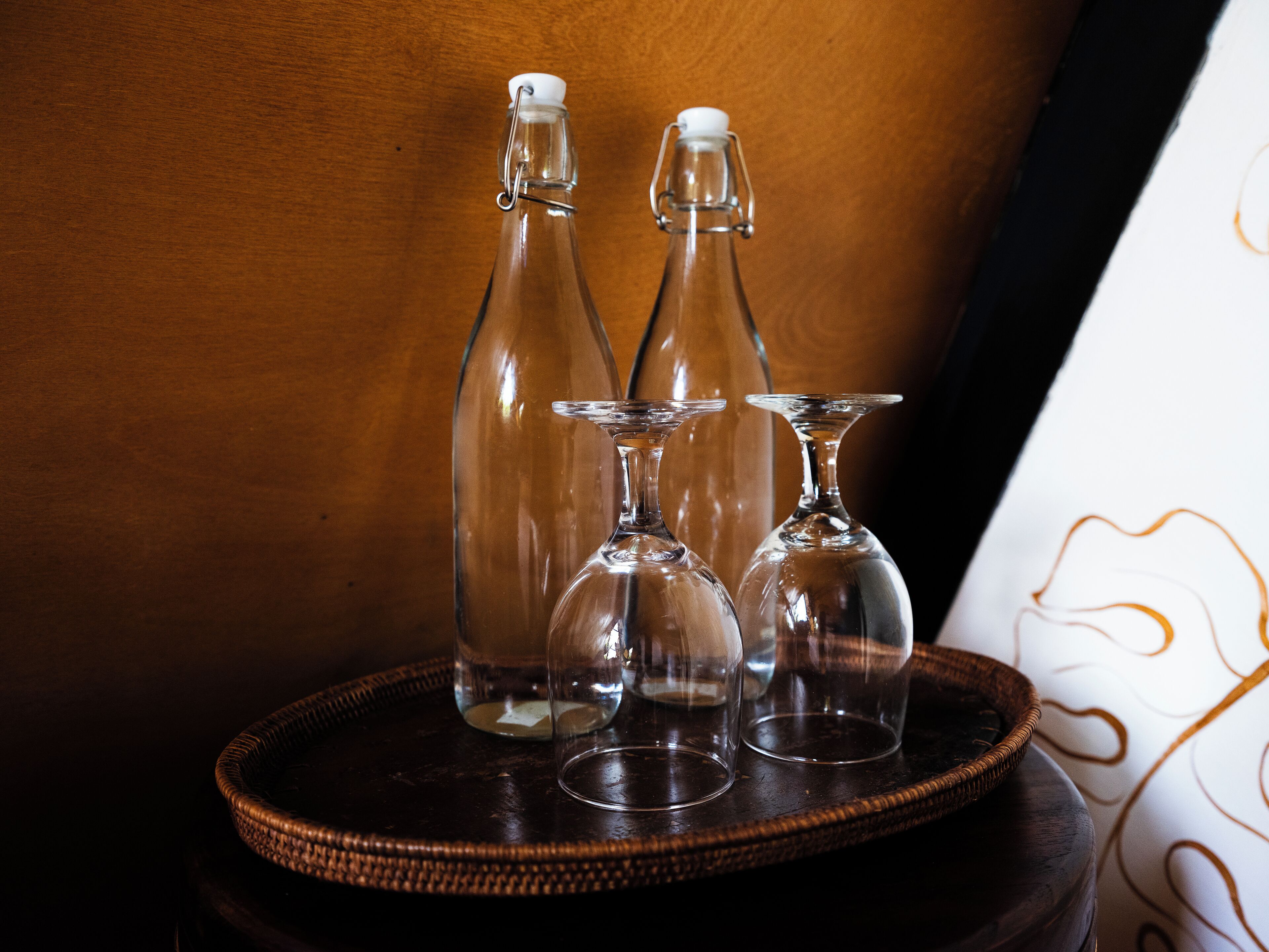 Una mesa de madera oscura con una bandeja tejida que contiene tres botellas de vidrio y dos copas de vino.