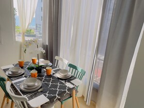 A dining area with a table set for four, chairs, and a view of the ocean through sheer curtains.