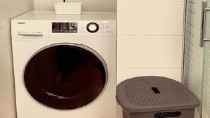 A compact, modern laundry room with a front-loading washer and dryer, a small trash bin, and a white tiled wall. - Apartment on the beach, new (Bat Yam)