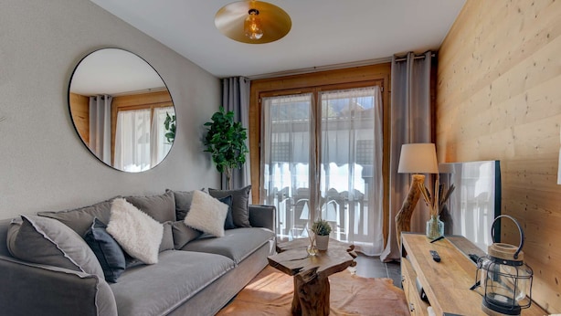 A cozy living room with a grey sofa, a round mirror, a wooden coffee table, and a wooden sideboard with a lamp and a lantern.