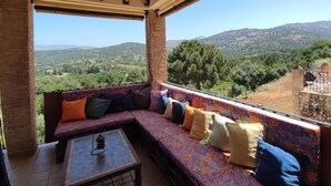 Een overdekt balkon met een bank en een tafel, met uitzicht op een glooiend landschap.
