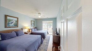 Ein Hotelzimmer mit zwei Betten, einem Schreibtisch, einem Stuhl, einem Deckenventilator und einem Fenster.