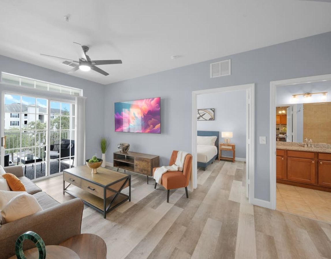 A spacious living room with a sofa, coffee table, and chairs, a dining area with a table and chairs, a kitchen with wooden cabinets, and a large window with a view of buildings.