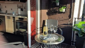 A balcony with a dining table, a bottle of wine, and two glasses. There is a kitchen area with a dishwasher and oven visible.