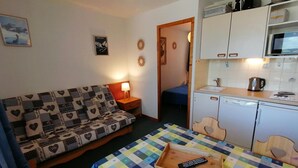 En kompakt opholdsstue med en sofa, et lille bord med en fjernbetjening, en mikrobølgeovn og et kompakt tekøkken.