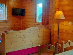 Quarto de madeira com uma cama, uma televisão presa na parede, uma janela com cortinas e um abajur.