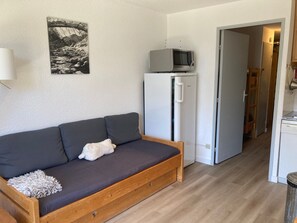 Un espace de vie compact comprenant un canapé, un réfrigérateur, un four à micro-ondes et une porte donnant sur un couloir.