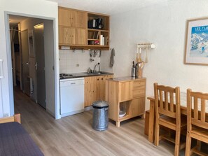 Cocina compacta con armarios de madera, un lavamanos y una estufa. Hay un cubo de basura y una pequeña mesa de madera con sillas.