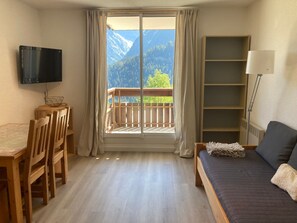 Una habitación con una mesa de comedor de madera, sillas, un sofá, un televisor, una estantería y un balcón con vistas a la montaña.