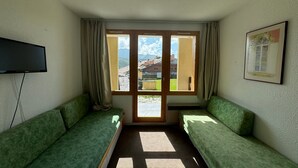 A room with two green sofas, a television on the wall, a window with curtains, and a framed picture. - Functional studio 3/4 pers with balcony and direct slopes access (Belle Plagne)