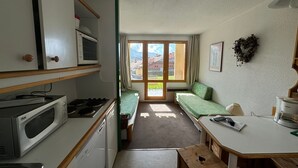 A compact kitchen with a stove, microwave, and a small refrigerator. There is a dining table with a bench, a green sofa, and a window with curtains. - Functional studio 3/4 pers with balcony and direct slopes access (Belle Plagne)