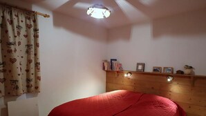 Ett sovrum med en sänggavel i trä, ett rött sängöverkast, ett element, en hylla med böcker och bilder samt en gardin med ett hjärtmönster.